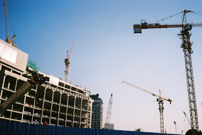 Multiple construction cranes are visible at a building site. A partially constructed building appears to the left, with cranes extended into the sky. The blue sky provides a clear backdrop, emphasizing the scale and complexity of the construction process.