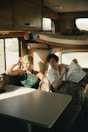 Two people are sitting inside a cozy and rustic caravan. The interior features wooden cabinets, a compact seating area with a table, and a window with a view of the outdoors. One person is holding a map, suggesting they are planning a journey or trip. The light streaming in through the window creates a warm and inviting atmosphere.