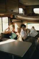 Two people are sitting inside a cozy and rustic caravan. The interior features wooden cabinets, a compact seating area with a table, and a window with a view of the outdoors. One person is holding a map, suggesting they are planning a journey or trip. The light streaming in through the window creates a warm and inviting atmosphere.