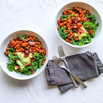 Two white bowls filled with a colorful salad featuring diced roasted sweet potatoes, leafy kale, crispy chickpeas, avocado slices, and bacon pieces. The bowls are placed on a textured white surface. A set of two silver forks is placed on top of a folded dark gray cloth napkin beside the dishes.