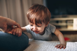 A young child with light brown hair is wearing a striped shirt and appears to be crying while reaching out toward an adult's hand. The background shows a blurred interior setting, possibly a living room.
