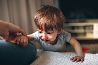A young child with light brown hair is wearing a striped shirt and appears to be crying while reaching out toward an adult's hand. The background shows a blurred interior setting, possibly a living room.