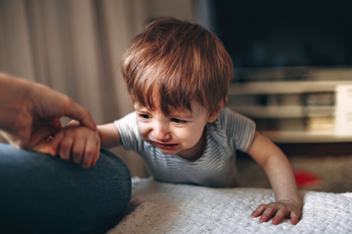 A young child with light brown hair is wearing a striped shirt and appears to be crying while reaching out toward an adult's hand. The background shows a blurred interior setting, possibly a living room.