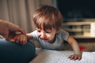 A young child with light brown hair is wearing a striped shirt and appears to be crying while reaching out toward an adult's hand. The background shows a blurred interior setting, possibly a living room.