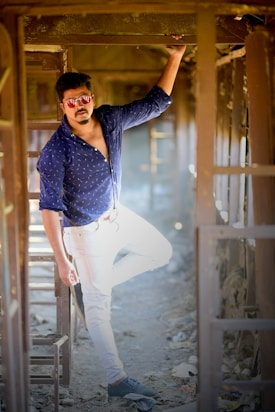 A man wearing a blue shirt and white pants stands confidently in an abandoned, rustic space. He is wearing reflective sunglasses and has one arm raised, resting against the structure. The lighting creates dramatic shadows and highlights, adding to the overall rugged and stylish look.