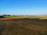 A vast agricultural field with a mixture of brown plowed soil and green crops in the distance. Irrigation pipes are laid out across the field. The skyline is clear with trees scattered along the horizon under a bright blue sky.