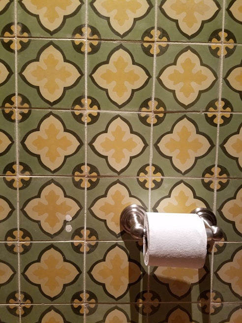 A roll of toilet paper is mounted on a holder that is attached to a wall with decorative green and yellow tiles. The tiles feature a repeating pattern of floral-like designs.