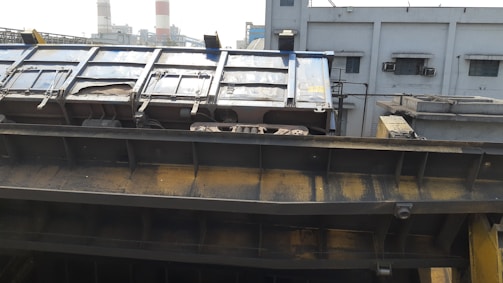 A large industrial structure appears to be part of a factory or processing plant. The metal surface has a worn, rusted appearance with visible bolts and mechanisms. Background buildings with small windows and several exhaust vents are visible, suggesting an industrial setting.