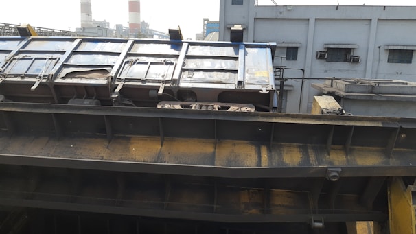 A large industrial structure appears to be part of a factory or processing plant. The metal surface has a worn, rusted appearance with visible bolts and mechanisms. Background buildings with small windows and several exhaust vents are visible, suggesting an industrial setting.