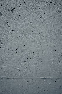A close-up view of a textured concrete wall featuring small holes and rough patches throughout the surface. The wall displays a uniform gray color with subtle shadows and highlights that emphasize its uneven texture.