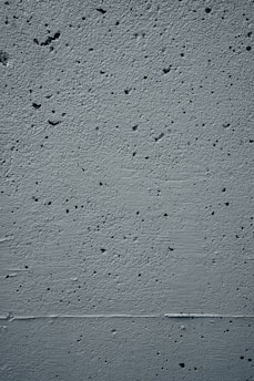 A close-up view of a textured concrete wall featuring small holes and rough patches throughout the surface. The wall displays a uniform gray color with subtle shadows and highlights that emphasize its uneven texture.