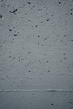 A close-up view of a textured concrete wall featuring small holes and rough patches throughout the surface. The wall displays a uniform gray color with subtle shadows and highlights that emphasize its uneven texture.