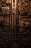 An intricate collection of gears and mechanical components arranged in a vintage, industrial style. The metal parts are rusted and have an antique appearance, adding to the overall gritty and rugged aesthetic.