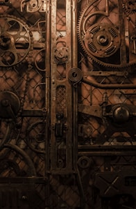 An intricate collection of gears and mechanical components arranged in a vintage, industrial style. The metal parts are rusted and have an antique appearance, adding to the overall gritty and rugged aesthetic.