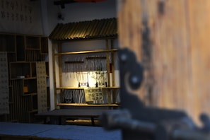 A room with traditional decor, featuring wooden furnishings and bamboo shelves. Calligraphy scrolls are displayed, and a small plant sits under focused lighting. The perspective is slightly obscured by a large wooden door on the right side.