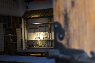 A room with traditional decor, featuring wooden furnishings and bamboo shelves. Calligraphy scrolls are displayed, and a small plant sits under focused lighting. The perspective is slightly obscured by a large wooden door on the right side.