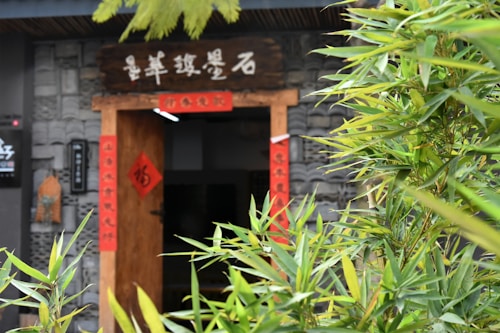 Entrance of a building decorated with traditional Chinese elements, including red scrolls with calligraphy on either side of a wooden door. Lush green bamboo plants are in the foreground, partially obscuring the view. The stone wall in the background is intricately designed, contributing to an authentic and cultural atmosphere.