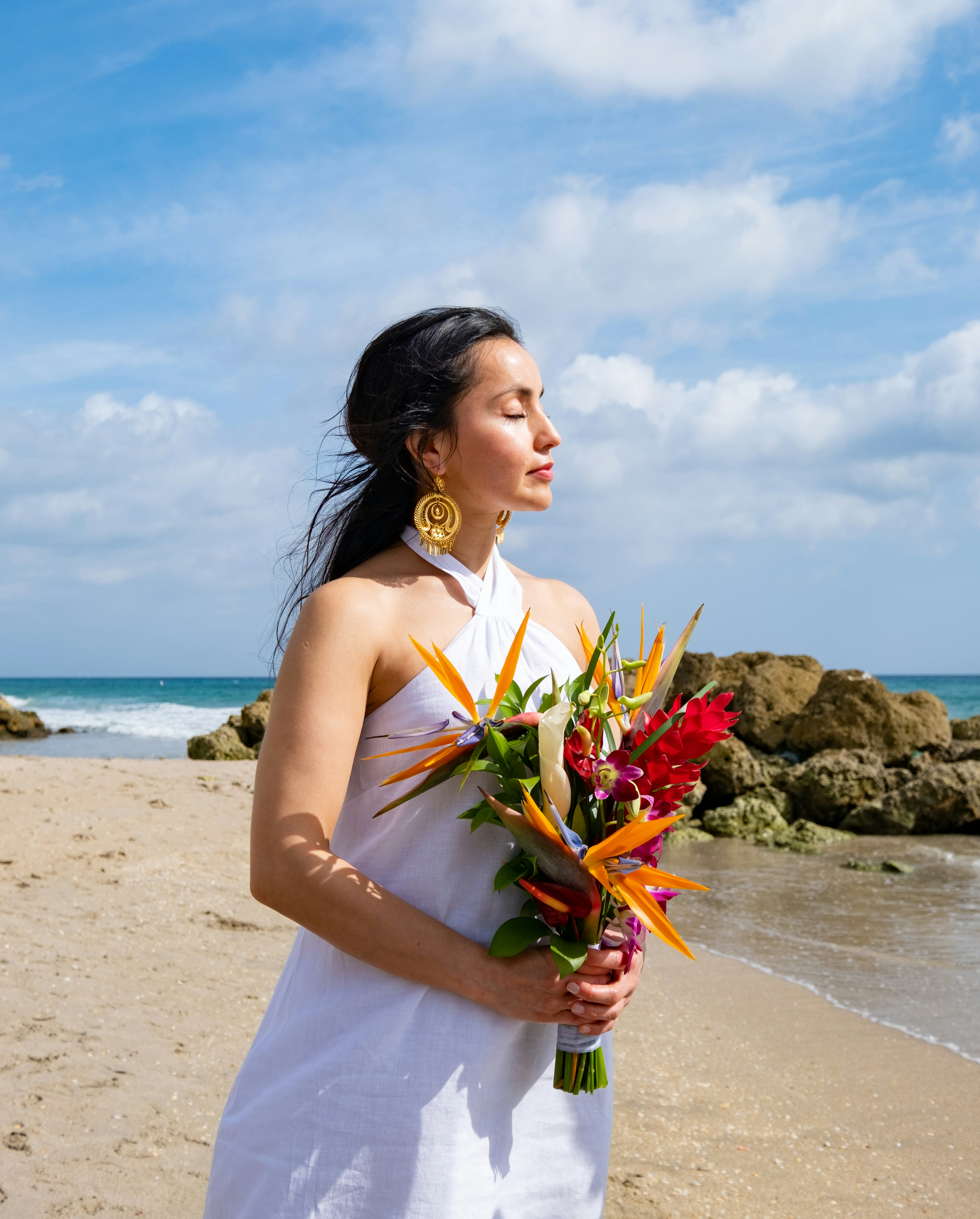 woman in white spaghetti strap dress holding bouquet of flowers standing on beach during daytime