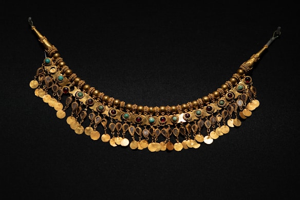 A decorative golden necklace featuring intricate beadwork and a series of hanging circular pendants. The necklace is adorned with round colorful gemstones alternating in turquoise and red, creating a striking contrast against the dark background.