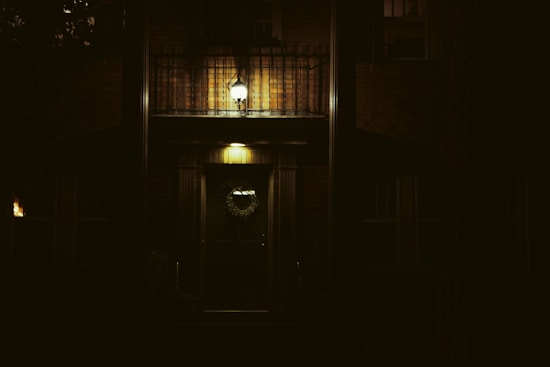 A dimly lit house entrance featuring a dark wooden door adorned with a wreath. Above the door, a small balcony with a metal railing is illuminated by a bright lamp. The surrounding area is in deep shadow, emphasizing the warm glow of the lights.