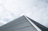 A metal roof with a corrugated texture ascends towards the sky, forming a sharp peak. Overhead, a blanket of soft clouds fills the background, providing a muted contrast to the structure.
