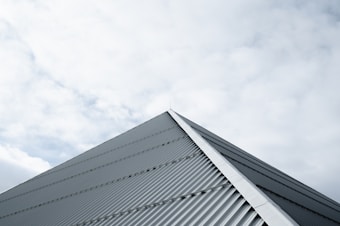 A metal roof with a corrugated texture ascends towards the sky, forming a sharp peak. Overhead, a blanket of soft clouds fills the background, providing a muted contrast to the structure.