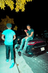 man in blue t-shirt and blue denim jeans standing beside black car during night time