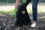 A person wearing green rubber gloves is picking up litter and placing it into a black bag that has a yellow label. The scene is outdoors near a body of water, with grass and leaves scattered on the ground.