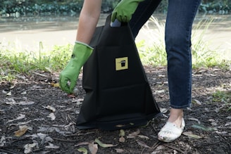 A person wearing green rubber gloves is picking up litter and placing it into a black bag that has a yellow label. The scene is outdoors near a body of water, with grass and leaves scattered on the ground.