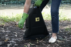A person wearing green rubber gloves is picking up litter and placing it into a black bag that has a yellow label. The scene is outdoors near a body of water, with grass and leaves scattered on the ground.