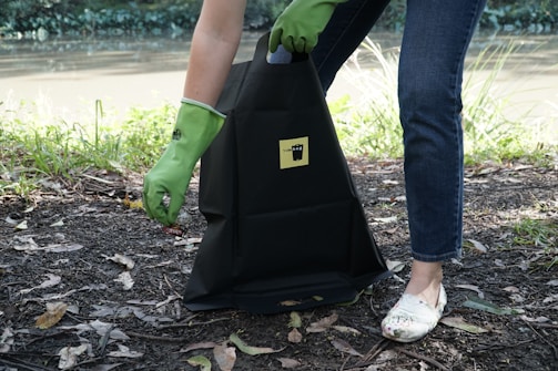 A person wearing green rubber gloves is picking up litter and placing it into a black bag that has a yellow label. The scene is outdoors near a body of water, with grass and leaves scattered on the ground.