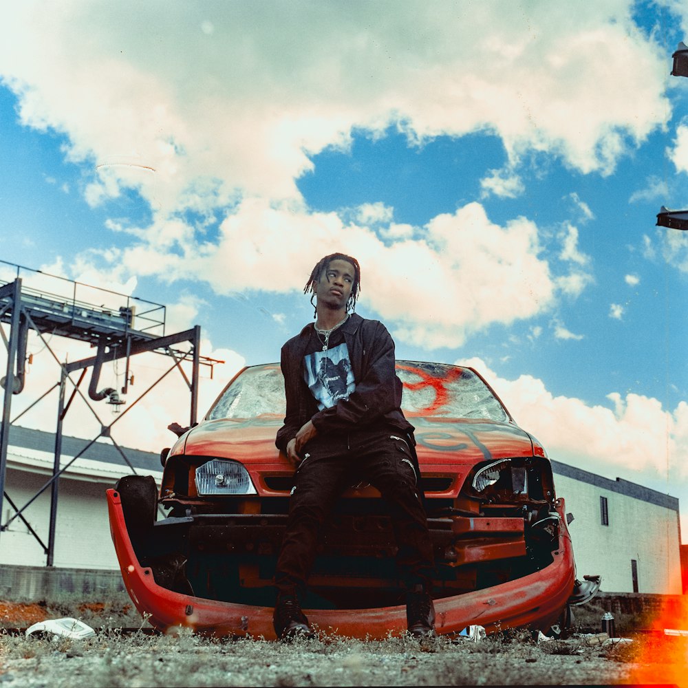 man in black jacket sitting on red car hood during daytime