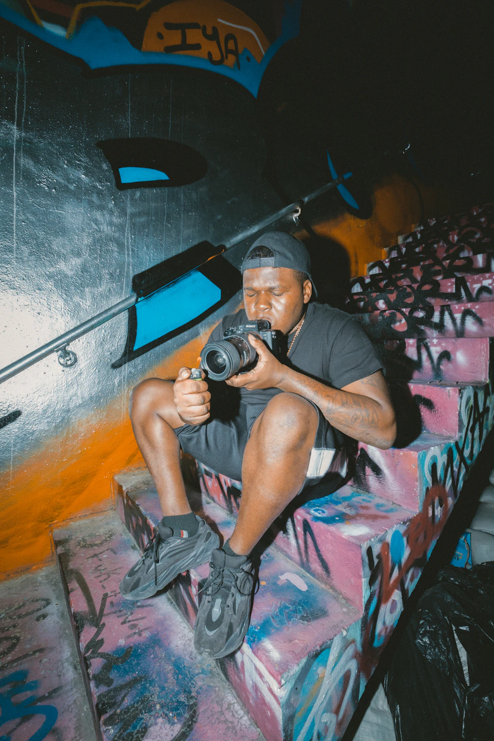 In the image, a young man is sitting on a set of stairs with his legs crossed and holding a camera up to his face. The stairs are covered in graffiti art, adding an urban feel to the scene. The man's attire consists of a black t-shirt and shorts, which contrasts with the colorful graffiti surrounding him.