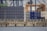 A large collection of stacked shipping containers, predominantly blue with some in green and white, is arranged along an industrial dockyard. Cranes and other port infrastructure are visible in the background. The dock is bordered by a concrete sea wall, with a narrow strip of land and some greenery visible between the wall and the containers. The water of the harbor is in the foreground.