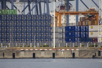 A large collection of stacked shipping containers, predominantly blue with some in green and white, is arranged along an industrial dockyard. Cranes and other port infrastructure are visible in the background. The dock is bordered by a concrete sea wall, with a narrow strip of land and some greenery visible between the wall and the containers. The water of the harbor is in the foreground.