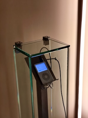 An iPod is encased in a transparent glass display stand, mounted on a metallic holder. Earphones are connected to the iPod, which displays a list on its screen.