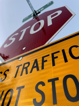 The image captures a close-up perspective of a stop sign and a traffic sign beneath it. The stop sign is red with white text, and the yellow traffic sign has black letters that are partially visible. A street sign can be seen at the top.