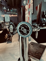 A cold brew dispenser with two taps is prominently displayed on a countertop. The labels on the taps indicate one is for Nitro Cold Brew and the other for regular Cold Brew. The background includes colorful decorations such as white and red balloons, with wall-mounted signage and a neon sign reading 'Coffee Togetherness.'