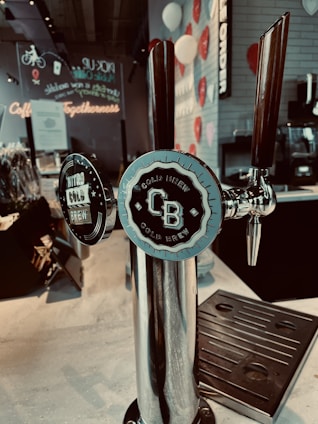 A cold brew dispenser with two taps is prominently displayed on a countertop. The labels on the taps indicate one is for Nitro Cold Brew and the other for regular Cold Brew. The background includes colorful decorations such as white and red balloons, with wall-mounted signage and a neon sign reading 'Coffee Togetherness.'