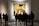A group of people stands in an art gallery observing a large framed painting. The painting appears to be a classical artwork with a complex scene involving numerous figures and intricate details. The gallery has a patterned floor and a softly lit ambiance.