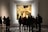 A group of people stands in an art gallery observing a large framed painting. The painting appears to be a classical artwork with a complex scene involving numerous figures and intricate details. The gallery has a patterned floor and a softly lit ambiance.