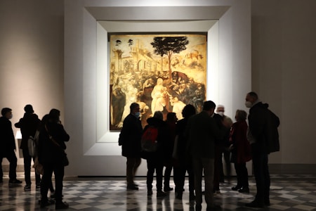A group of people stands in an art gallery observing a large framed painting. The painting appears to be a classical artwork with a complex scene involving numerous figures and intricate details. The gallery has a patterned floor and a softly lit ambiance.