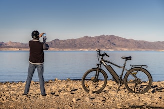man in black jacket and gray pants standing beside black bicycle near body of water during