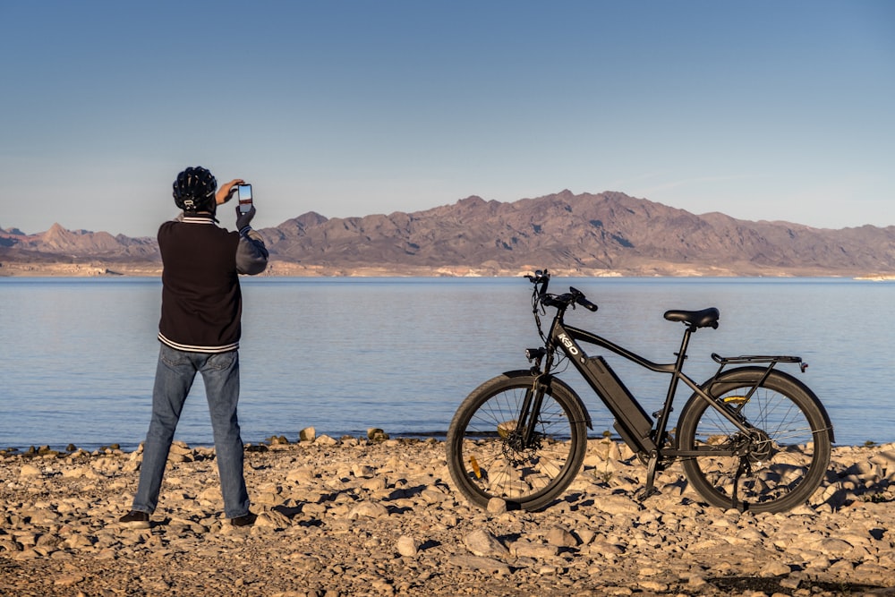 man in black jacket and gray pants standing beside black bicycle near body of water during
