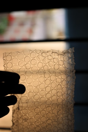 A translucent sheet of bubble wrap is held up against a blurred background, with light filtering through it, illuminating the bubble pattern. A hand on the left side holds the bubble wrap, providing a silhouette contrast against the lighter background.