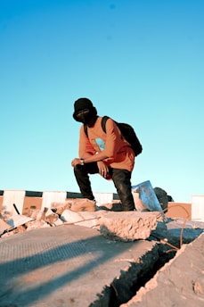 A person wearing a mask and orange shirt crouches on a pile of rubble under a clear blue sky. The individual is wearing dark pants and boots, with a backpack on their back. Sunlight casts shadows across the debris.