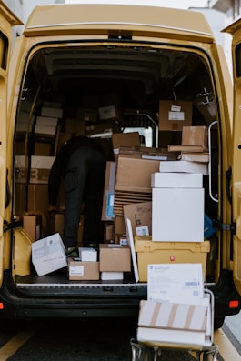 A delivery van with its back doors open, packed with numerous cardboard boxes of various sizes. A person is partially visible inside the van, surrounded by the boxes. The scene suggests the process of loading or unloading packages.