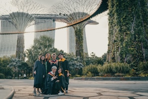 A group of six people in graduation attire are celebrating outdoors with greenery and a distinctive architectural structure in the background. The graduates are smiling, with some making peace signs, and the atmosphere suggests a sense of achievement and joy.