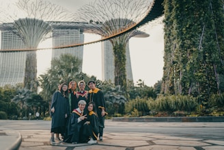 A group of six people in graduation attire are celebrating outdoors with greenery and a distinctive architectural structure in the background. The graduates are smiling, with some making peace signs, and the atmosphere suggests a sense of achievement and joy.