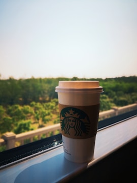 A paper cup with a recognizable green logo sits on a ledge by a window, overlooking a lush green landscape under a clear sky.